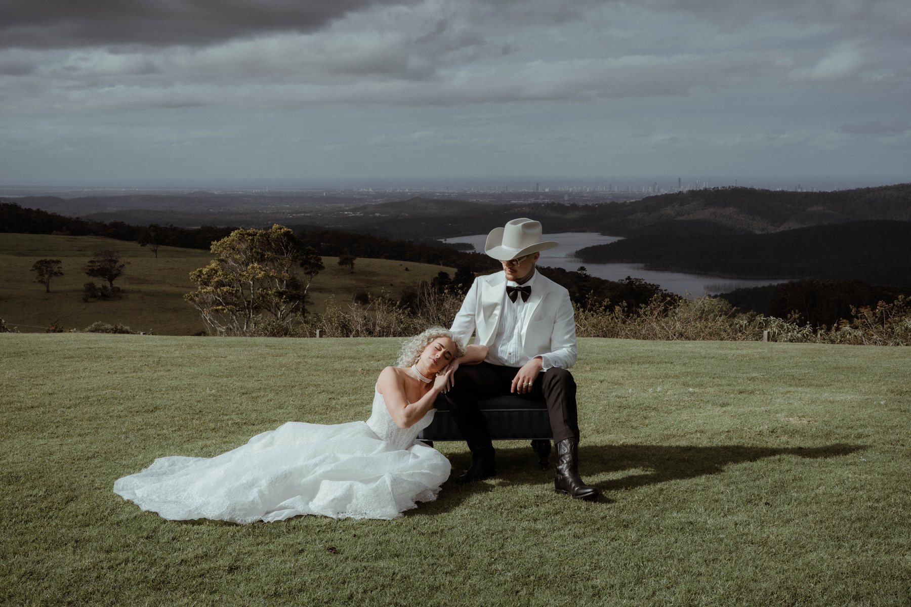 Kaleb seated on the grass with Monique, the ranges behind, at Rosewood Estate, Beechmont QLD — wedding photography by C2 Films